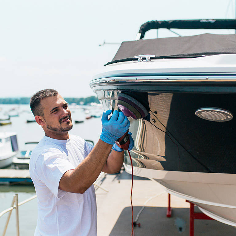 A technician polishing the hull of a boat at a marina, using a power tool to ensure the vessel's surface is well-maintained, with various boats visible in the background.