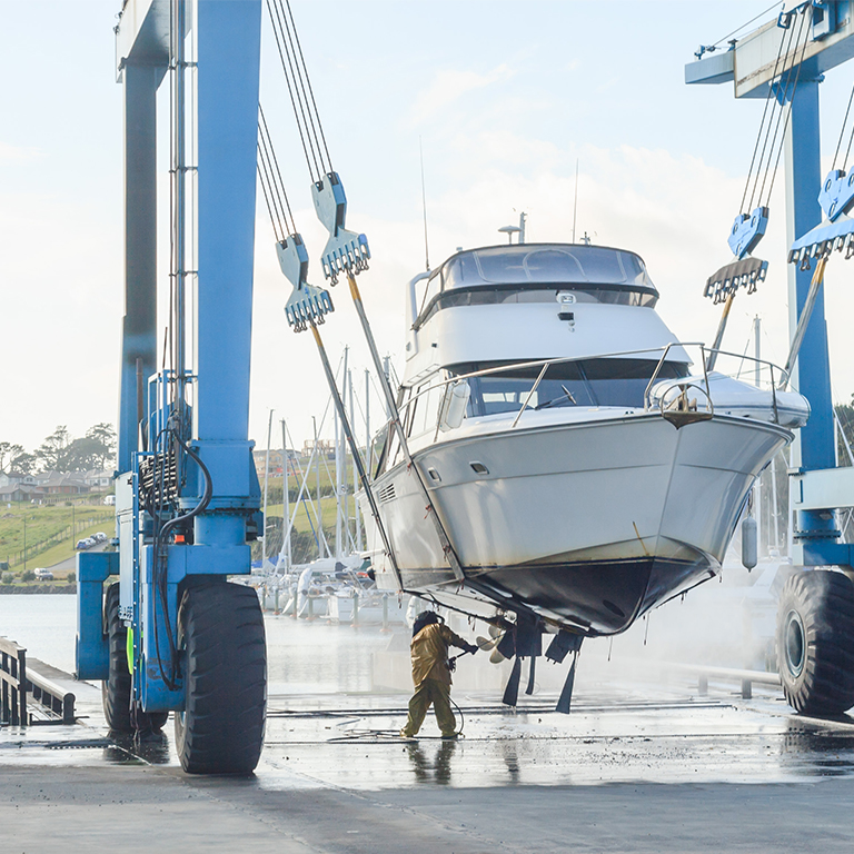 A person performing boat maintenance by cleaning the hull of a yacht suspended by a large blue boat lift at a marina.