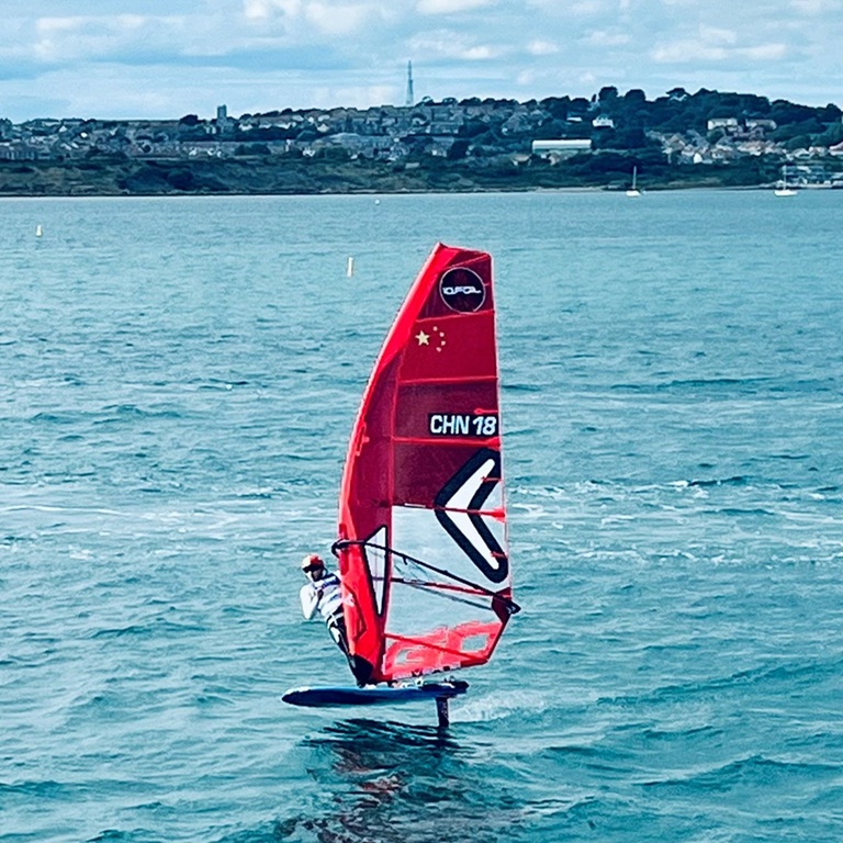 iQFOiL windsurfer riding above the water on a hydrofoil board, with a red sail and shoreline visible in the background.
