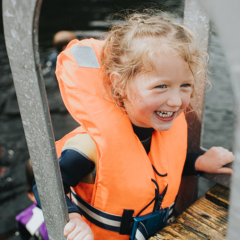 Happy and confident pre-school age girl climbing up a ladder onto a jetty wearing a fluorescent orange life jacket. Another child swims in the background.