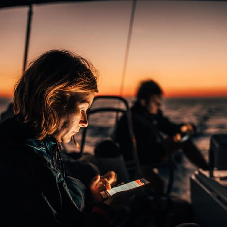 Two people on a boat at sunset using mobile devices, highlight the use of modern sailing equipment and technology for navigation and communication.