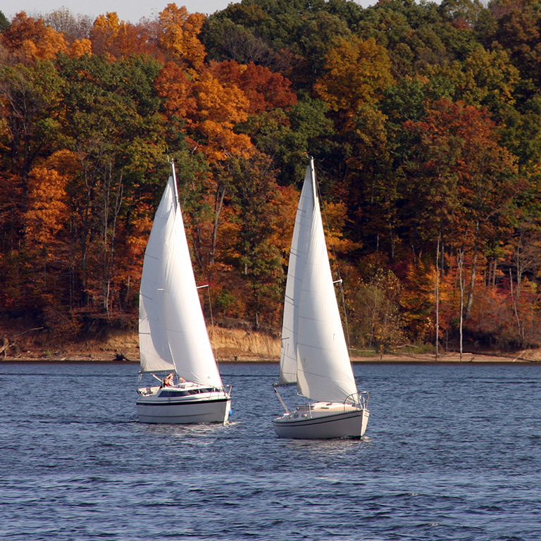 Two white sailboats sailing on a lake with autumn trees in shades of orange, yellow, and green in the background.