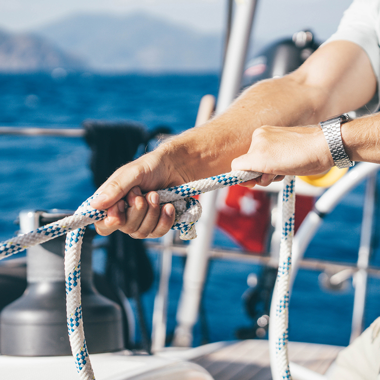 Hands holding a blue-and-white braided rope on a sailboat, with forearms and a wristwatch visible, ocean and distant shoreline in the background.