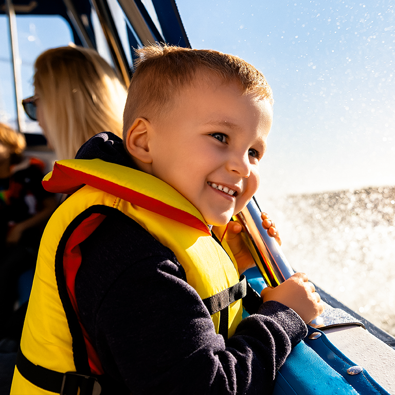 A child on a boat, wearing a life jacket looking out at the water spray on a sunny day.